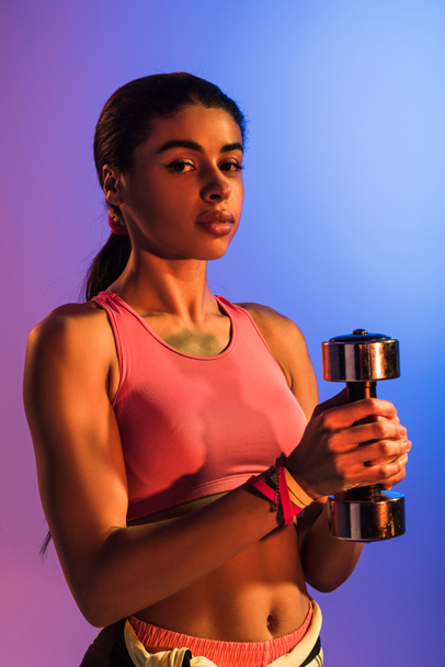 sportive african american girl holding dumbbell and looking at camera on purple and blue gradient background - Photo, Image