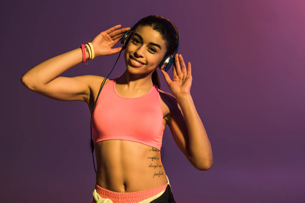 deportivo africano americano chica en rosa deportes sujetador y auriculares sonriendo y mirando a la cámara en púrpura fondo
 - Foto, imagen