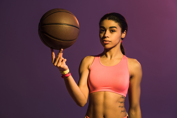 pretty african american girl in pink sports bra holding brown ball and looking at camera on purple background - Фото, изображение