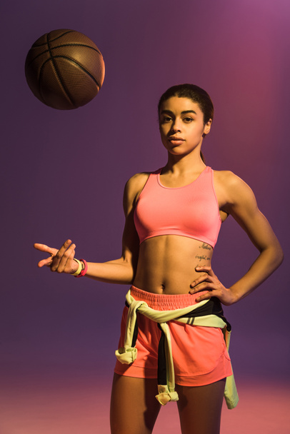 紫色の背景にカメラを見て茶色のボールを持つ美しいスポーティなアフリカ系アメリカ人の女の子 - 写真・画像