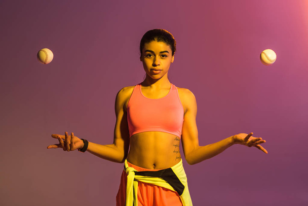 attractive african american girl in pink sports bra and shorts juggle with balls on purple background with gradient - Photo, Image