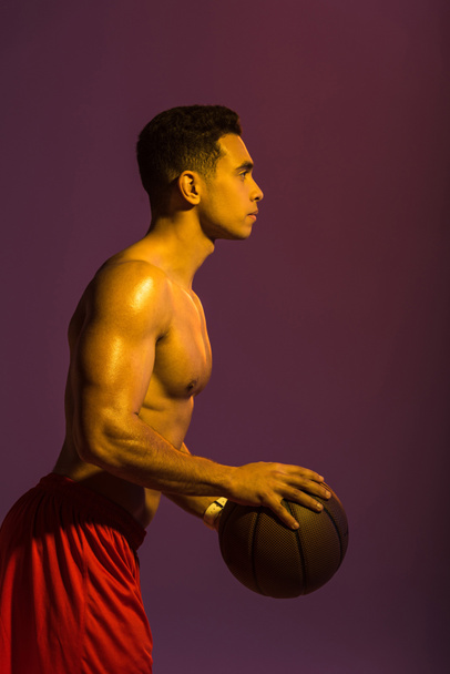 sportive shirtless mixed race man with muscular torso holding brown ball on purple background - Foto, Imagem