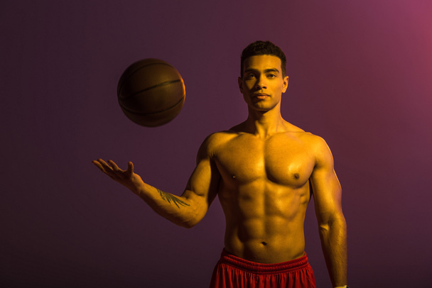 good-looking mixed race man with muscular torso playing ball on purple background - Photo, Image