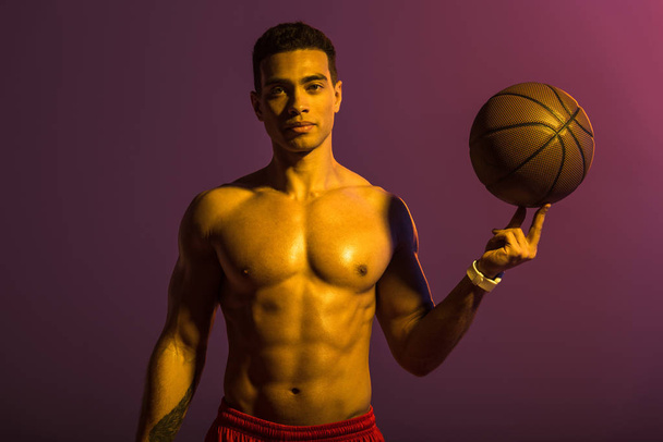 good-looking sportive mixed race man holding brown ball and looking at camera on purple background - Photo, Image