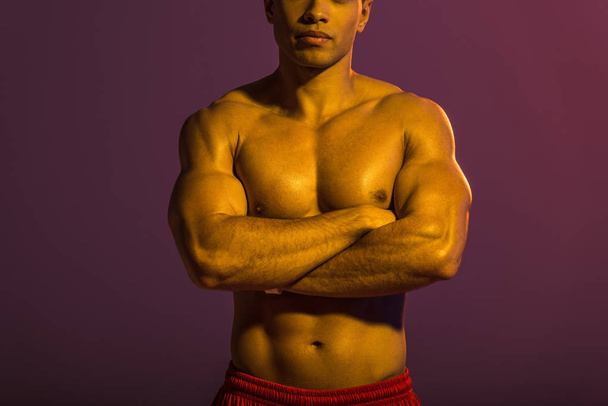 bijgesneden weergave van atletische man poseren op camera met gekruiste armen op paarse achtergrond - Foto, afbeelding
