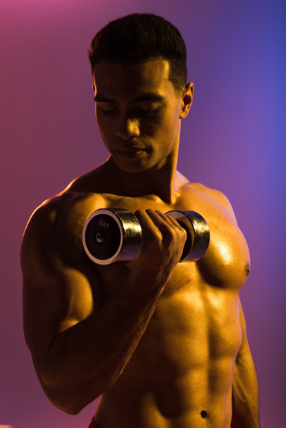 handsome sportive mixed race man training with dumbbell on purple and blue gradient background - Foto, immagini