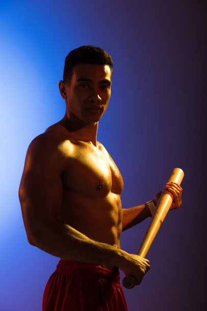 knap shirtless gemengde race man met honkbalknuppel en kijken naar camera op blauwe en donker paarse gradiënt achtergrond - Foto, afbeelding