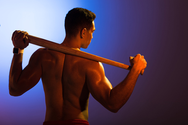 Achteraanzicht van shirtless sportieve mixed race man met honkbalknuppel op blauwe en donker paarse gradiënt achtergrond - Foto, afbeelding