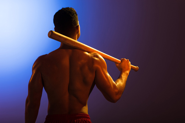 pohled na atletického smíšené muže s baseballovou pálkou na modrém a tmavém purpurovém pozadí - Fotografie, Obrázek