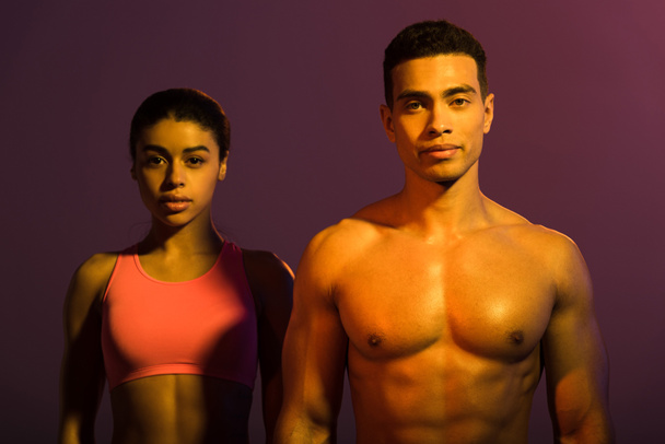 attractive african american woman in pink sports bra and shirtless mixed race man looking at camera on purple background - Fotografie, Obrázek