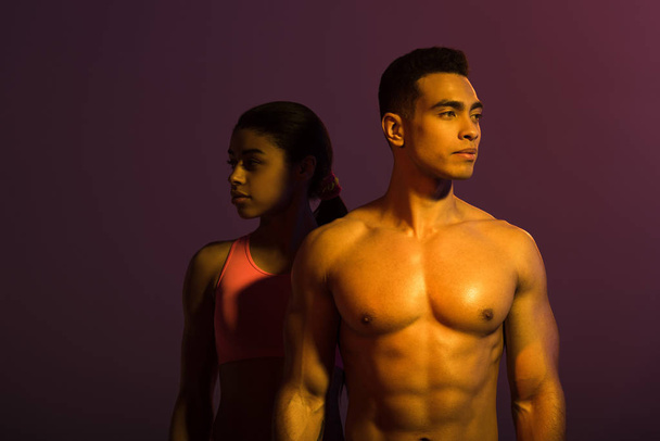 pretty african american woman in pink sports bra and shirtless mixed race man looking at camera on purple background - Φωτογραφία, εικόνα