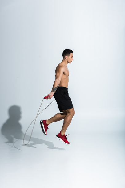 side view of mixed race man in black shorts and red sneakers jumping with skipping rope on white - Photo, Image