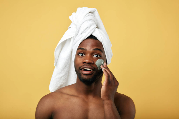 Young happy man with a towel on his head applying mask on his face - Photo, Image