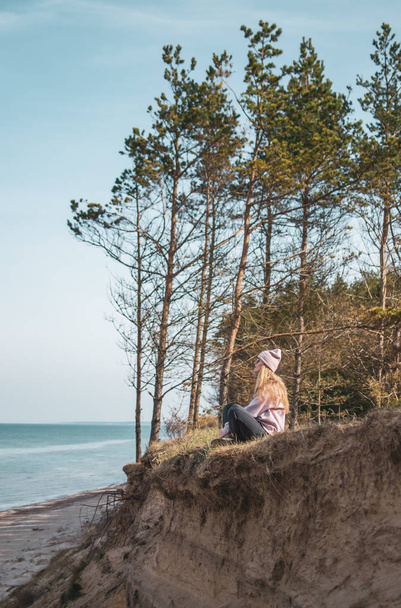 Young adult woman in pink hat sitting alone on the bluffs, looking of sea, freedom concept, peaceful atmosphere, meditation, copy space, Latvia, Jurkalne - Foto, Imagen