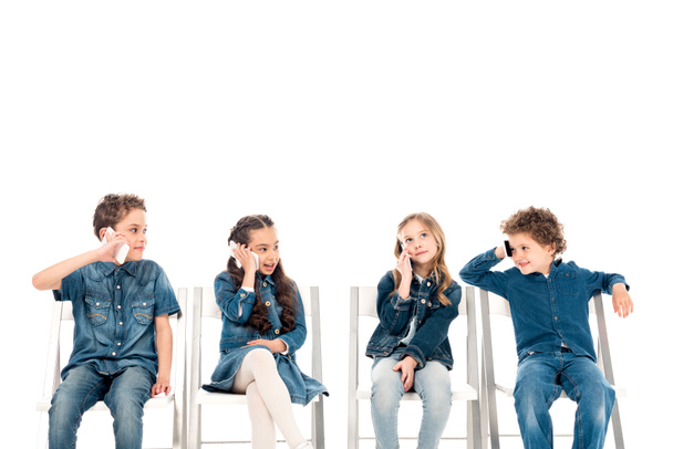 four kids in denim clothes sitting on chairs and talking on smartphones isolated on white - Photo, Image