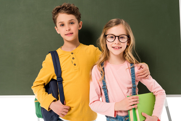 two schoolchildren with backpacks and books standing near blackboard isolated on white - Foto, Imagen