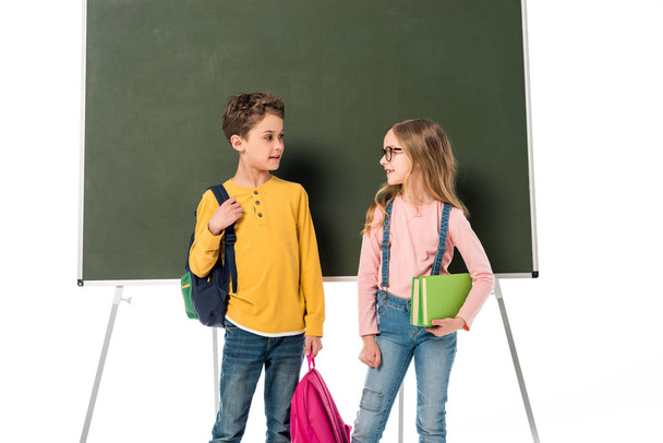 two schoolchildren with backpacks and books standing near blackboard isolated on white - Photo, Image