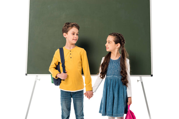 two schoolchildren with backpacks holding hands near blackboard isolated on white - Фото, изображение