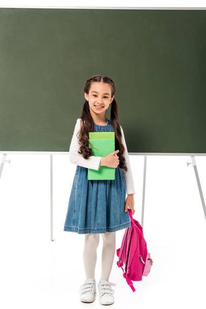 full length view of smiling schoolgirl with backpack and books standing near blackboard isolated on white - Photo, Image