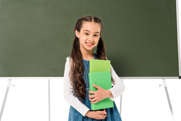 smiling schoolgirl holding books near blackboard isolated on white - Фото, изображение
