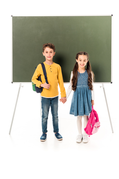full length view of two schoolchildren with backpacks holding hands near blackboard isolated on white - Foto, Imagen