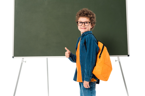 χαμογελαστός μαθητής με γυαλιά να στέκεται κοντά στον μαυροπίνακα και να δείχνει τον αντίχειρά του απομονωμένο σε λευκό - Φωτογραφία, εικόνα