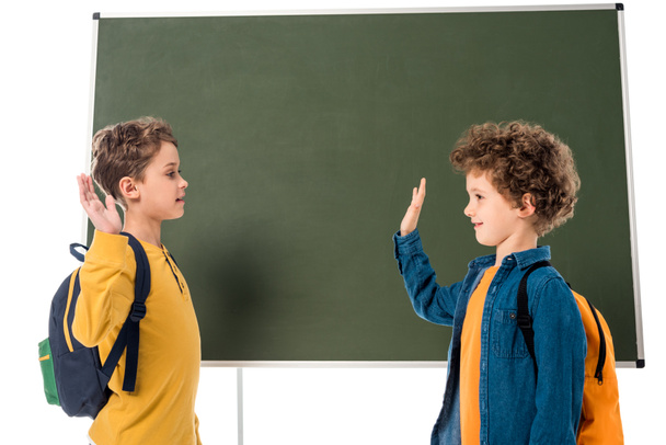 two smiling schoolboys with backpacks showing high five sign near blackboard isolated on white - Foto, immagini