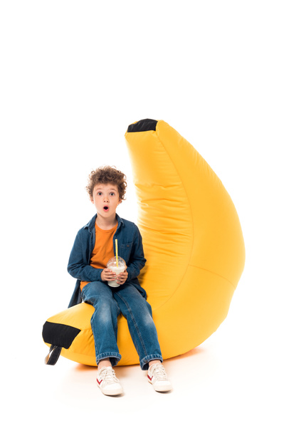 shocked child in jeans sitting on bean bag chair and holding milkshake on white - Photo, Image