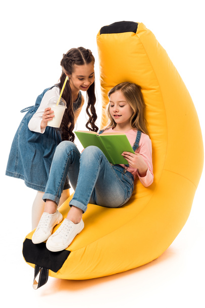 two smiling kids with bean bag chair reading book on white - Foto, afbeelding