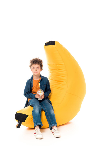 Kid in Casual kleding zittend op Bean Bag Chair en het houden van milkshake op wit - Foto, afbeelding