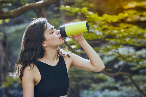 Здорова фітнес-дівчина п'є воду з зеленої пляшки в лісі
 - Фото, зображення
