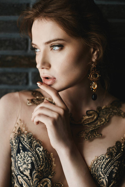 Beautiful, sexy, fashionable brunette girl with blue eyes, in gold dress with handmade gold earrings, is posing at brick wall background - Photo, Image