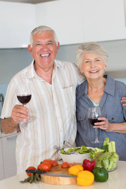 Smiling senior couple preparing a meal and having red wine at home in the kitchen - Photo, Image