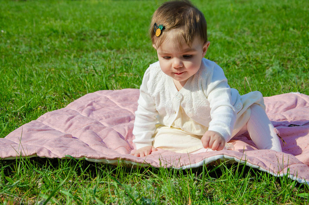 Baby in bright clothes on a pink plaid on green grass in the park. Newborn baby outside on a warm day. Little girl plays on a plaid on a green lawn in spring - Photo, Image