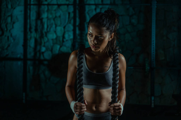 Σέξι μελαχρινή γυμναστήριο υγρή γυναίκα στο γυμναστήριο. Πορτραίτο του ελκυστικού κοριτσιού που παίρνει σπάσιμο μετά την άσκηση με σχοινιά μάχης - Φωτογραφία, εικόνα