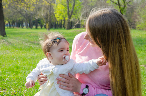 Mãe com bebê em roupas brilhantes em uma xadrez rosa na direita verde. Família descansando no parque em um dia quente. Mãe e menina 10 meses a pé no parque
 - Foto, Imagem