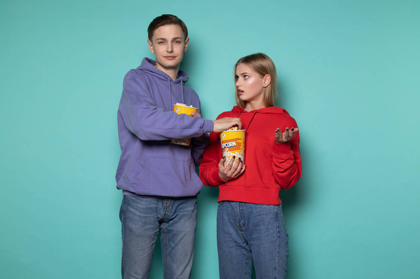 Young attractive man in blue hoodie stealing popcorn from his girlfriend, beautiful blonde girl looks at him indignantly - Photo, Image