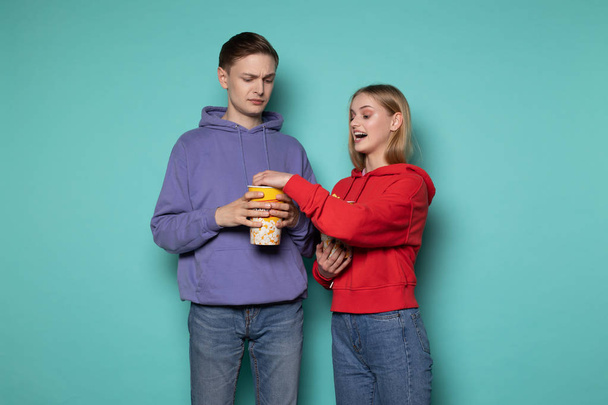 Beautiful blonde girl in red hoodie stealing popcorn from her boyfriend, young man looking at her indignantly - Photo, Image