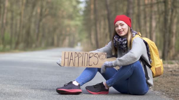 Szczęśliwy Autostopem dziewczyna siedzieć na pustej drodze w drewnie z plakatu szczęścia - Materiał filmowy, wideo