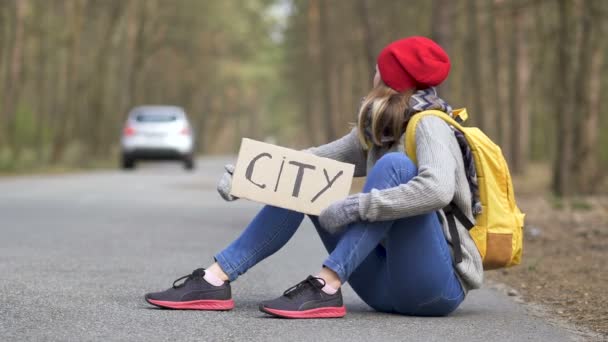 Dziewczyna autostopem siedzieć na pustej drodze w drewnie z plakatu miasta - Materiał filmowy, wideo
