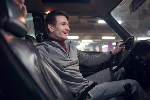 Εικόνα από την πλευρά του νεαρού χαμογελαστή άντρα κάθεται στο αυτοκίνητο. - Φωτογραφία, εικόνα