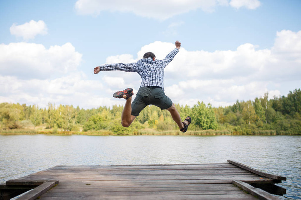 Photo from back of young jumping man on wooden bridge by river - Фото, изображение