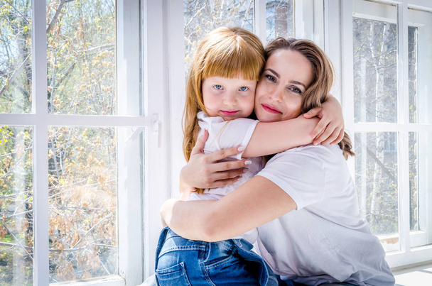 Mom hugs her little daughter with red hair sitting on the window - Фото, изображение