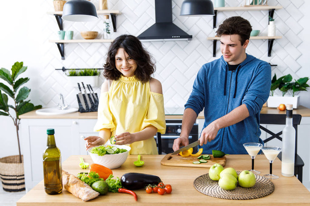 若いかわいい笑顔のカップルは自宅のキッチンで一緒に調理します。若い人たちは良いキッチンでサラダを作っています - 写真・画像