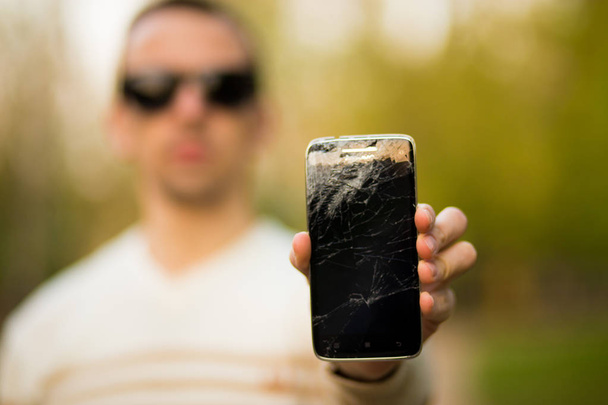 Guy houdt zwarte smartphone met gebroken display. Gebroken scherm van moderne frameloze telefoon. - Foto, afbeelding