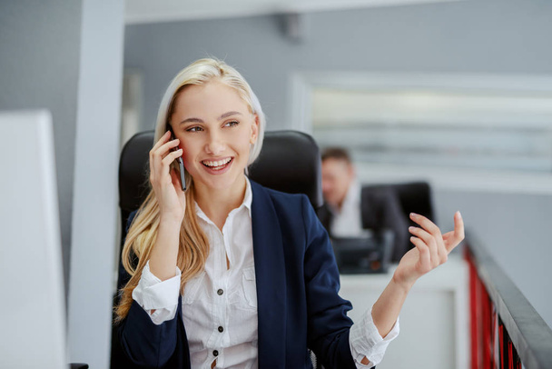 Giovane bionda sorridente donna d'affari caucasica seduta in ufficio e che utilizza lo smart phone per parlare d'affari
.  - Foto, immagini