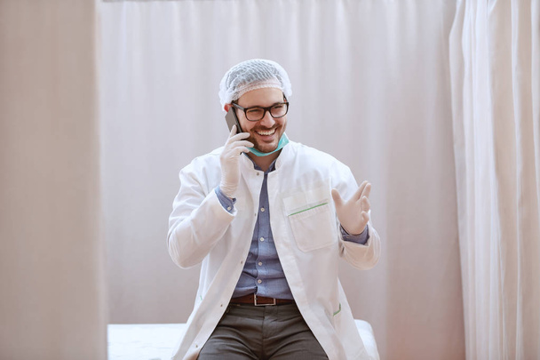 Usmívající se Kavkazský chirurg, který sedí na nemocniční posteli a vykládá dobré zprávy přes chytrého telefonu. - Fotografie, Obrázek