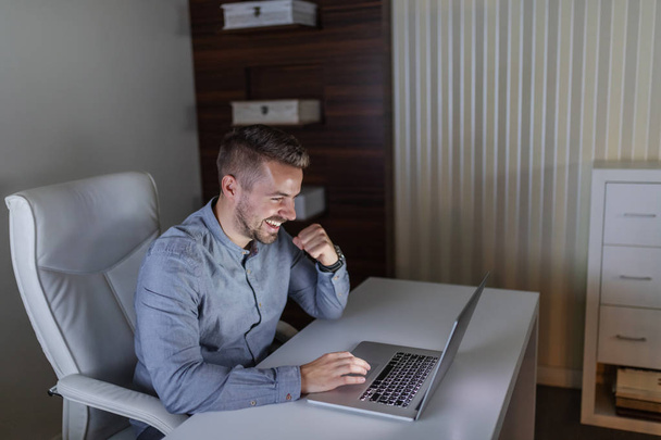 Hymyilevä komea valkoihoinen freelancer istuu toimistossa myöhään illalla ja työskentelee kannettavan tietokoneen
. - Valokuva, kuva