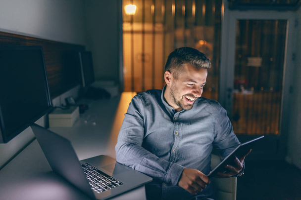 Καυκάσιος όμορφος γενειοφόρος freelancer με toothy χαμόγελο, κάθεται στο γραφείο αργά τη νύχτα και χρήση ταμπλετών. Δίπλα του στο γραφείο laptop. - Φωτογραφία, εικόνα