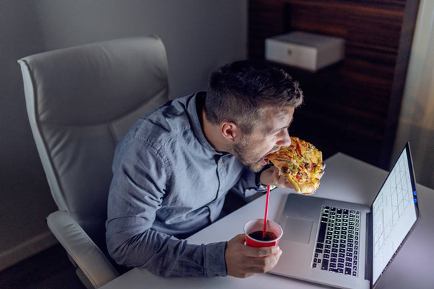 Young Caucasian architect eating pizza and drinking juice at office. On desk laptop. Late night work concept. - Photo, Image